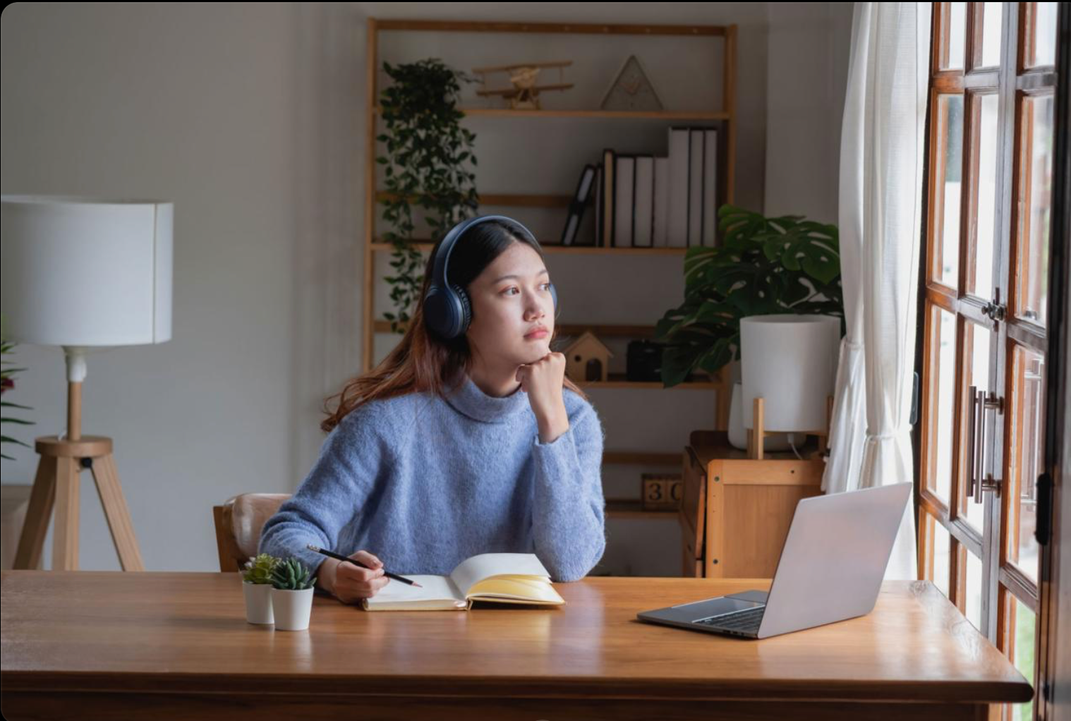 Mujer con audífonos reflexionando junto a una ventana, con un cuaderno abierto